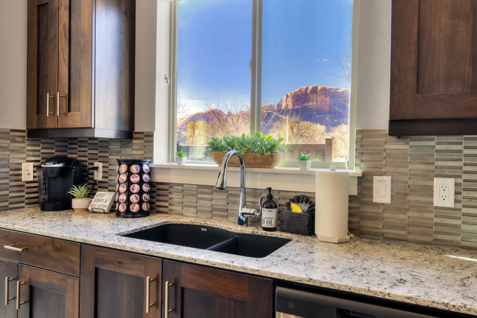 Sunlit Modern Kitchen: Dark Wood, Granite Counter, and Panoramic Window View A clean kitchen layout with a carousel of pods for the Keurig. Drip coffee maker also available in this Entrada Moab townhome.
